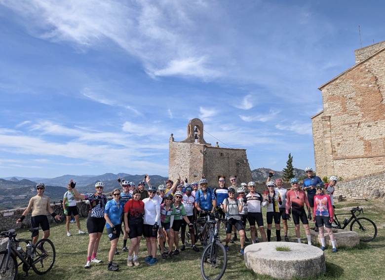A large group of cyclists posing for a photo in front of a historic building and bell tower, with mountains and a blue sky in the background.