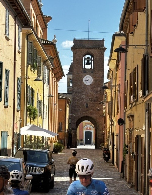 A narrow cobblestone street lined with colorful buildings, leading to a clock tower in the distance. Cyclists and pedestrians are visible in the foreground, with an awning-covered table on the left.