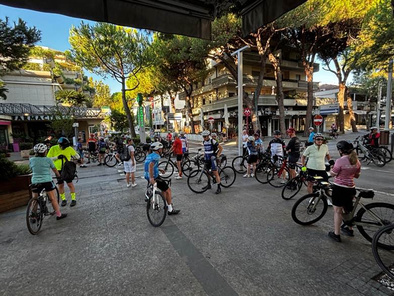 A group of cyclists gathered on a street, waiting to start their ride. Trees are lining the road, and buildings are visible in the background.
