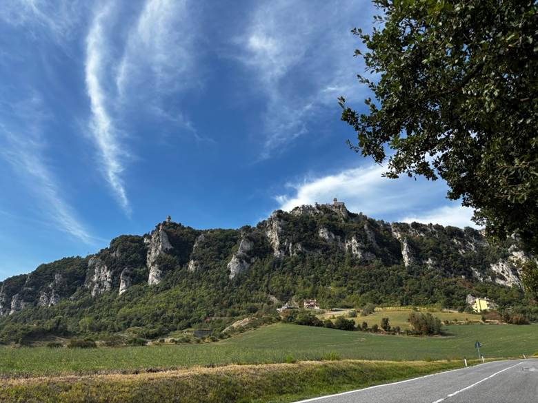 Scenic view of a rocky hillside with lush greenery and a blue sky with clouds. A road runs in the foreground.
