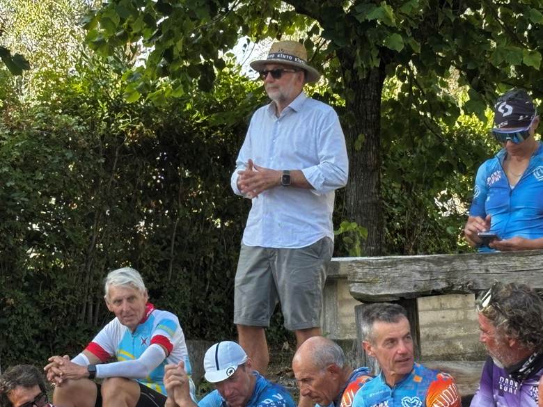 A group of cyclists sitting and standing outdoors, with one man wearing a straw hat and standing with his hands clasped while others are seated casually, some in cycling gear.