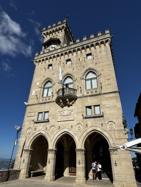 A tall historic building with a turret and gothic architectural features, set against a clear blue sky. The building displays arched windows and a balcony, with people standing near the entrance.
