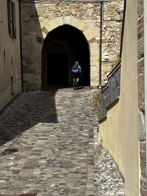 A cyclist riding up a cobblestone path towards a stone archway in a historic area, surrounded by old buildings.