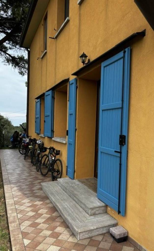 A charming yellow house with blue shutters and a welcoming entrance, featuring steps leading up to the doorway. Bicycles are parked along the side on a patterned walkway.