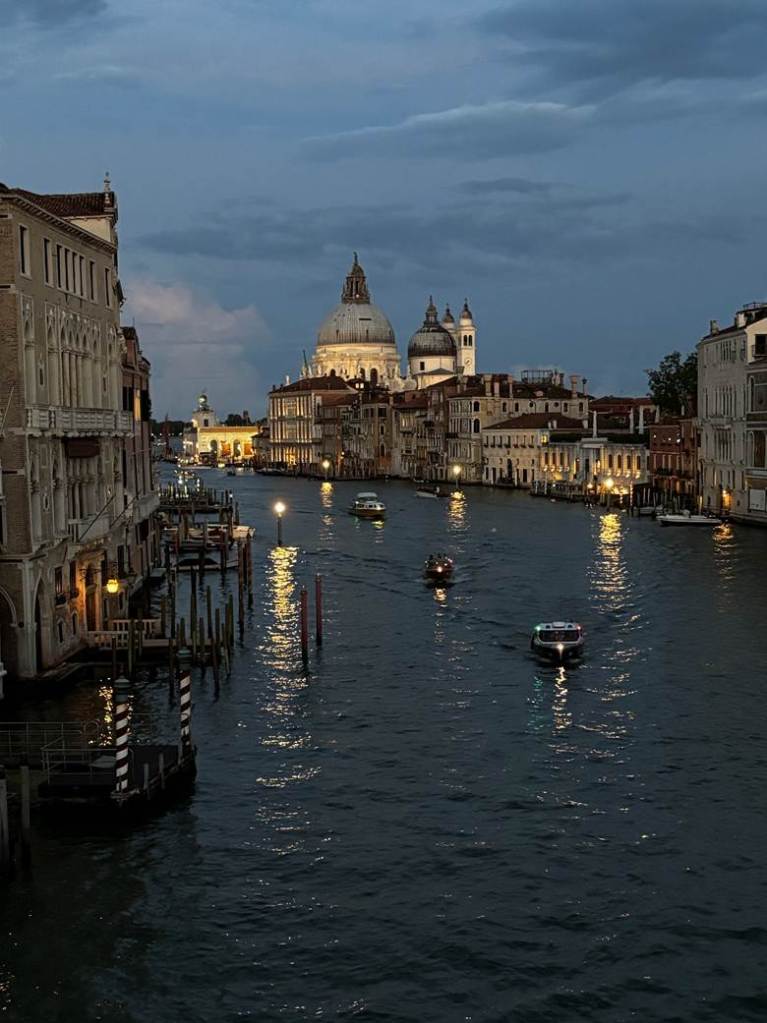 A view of a canal in Venice at dusk, featuring illuminated buildings and boats on the water, with a prominent dome structure in the background.