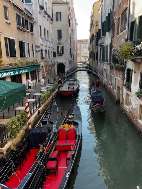 A scenic canal in Venice featuring gondolas and small boats, flanked by historic buildings and a restaurant with outdoor seating.
