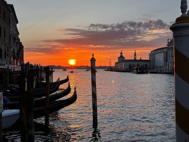 Sunset over a canal in Venice, with gondolas lined up along the shore and buildings reflected in the water.